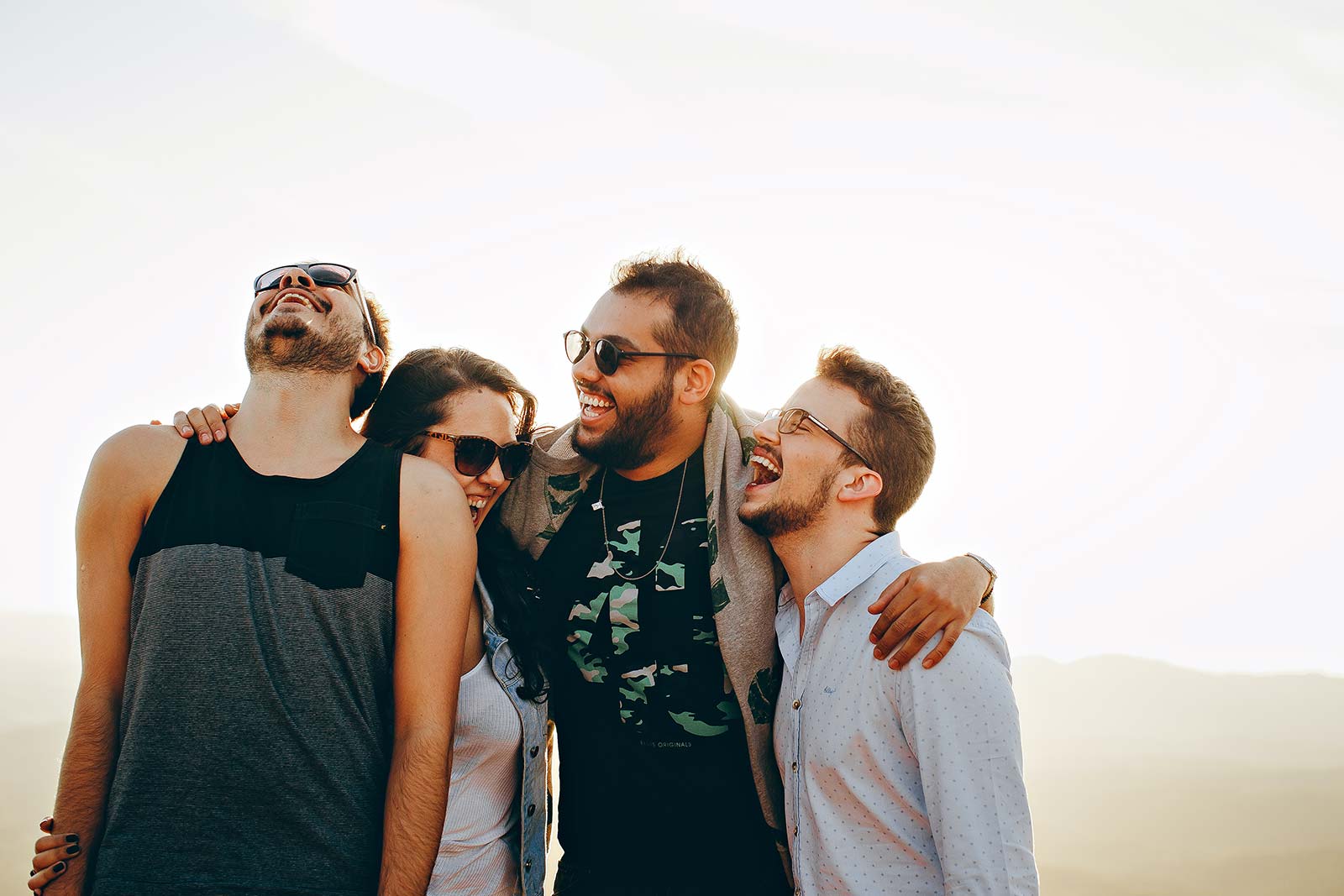 Four friends stand closely together outdoors, smiling and laughing in the sun. Dressed casually, two wear sunglasses, enjoying a warm atmosphere—just like you can with Stay Now Pay Later at Cape Town hotels for your next getaway.